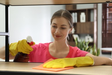 Young woman cleaning wooden shelf with rag and spray in office