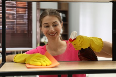 Photo of Young woman cleaning wooden shelf with rag and spray in office