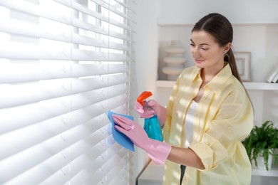 Young woman cleaning window blinds with rag and spray at home