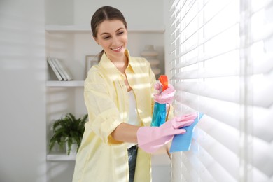 Young woman cleaning window blinds with rag and spray at home