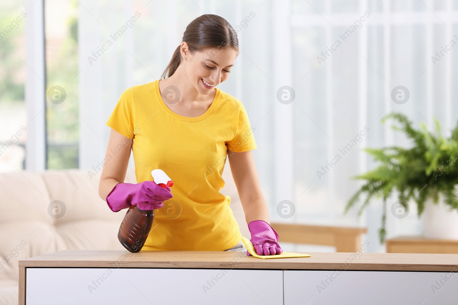 Photo of Young woman cleaning wooden table with rag and spray at home