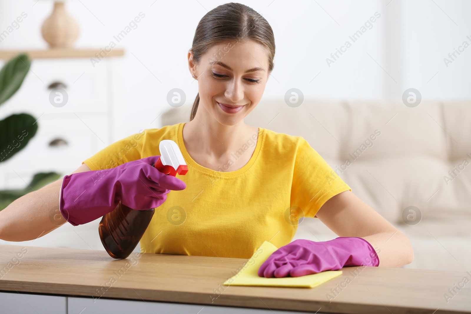 Photo of Young woman cleaning wooden table with rag and spray at home