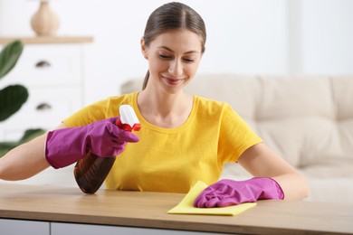 Photo of Young woman cleaning wooden table with rag and spray at home