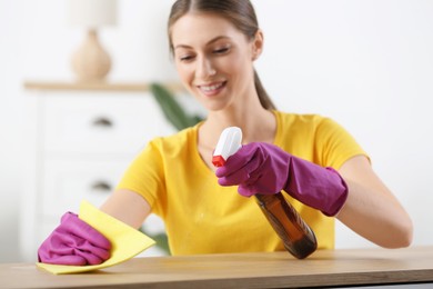 Young woman cleaning wooden table with rag and spray at home, selective focus