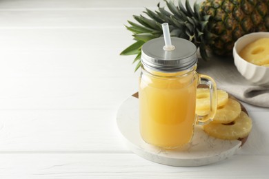 Photo of Tasty pineapple juice in mason jar and slices of fresh fruit on white wooden table, closeup. Space for text