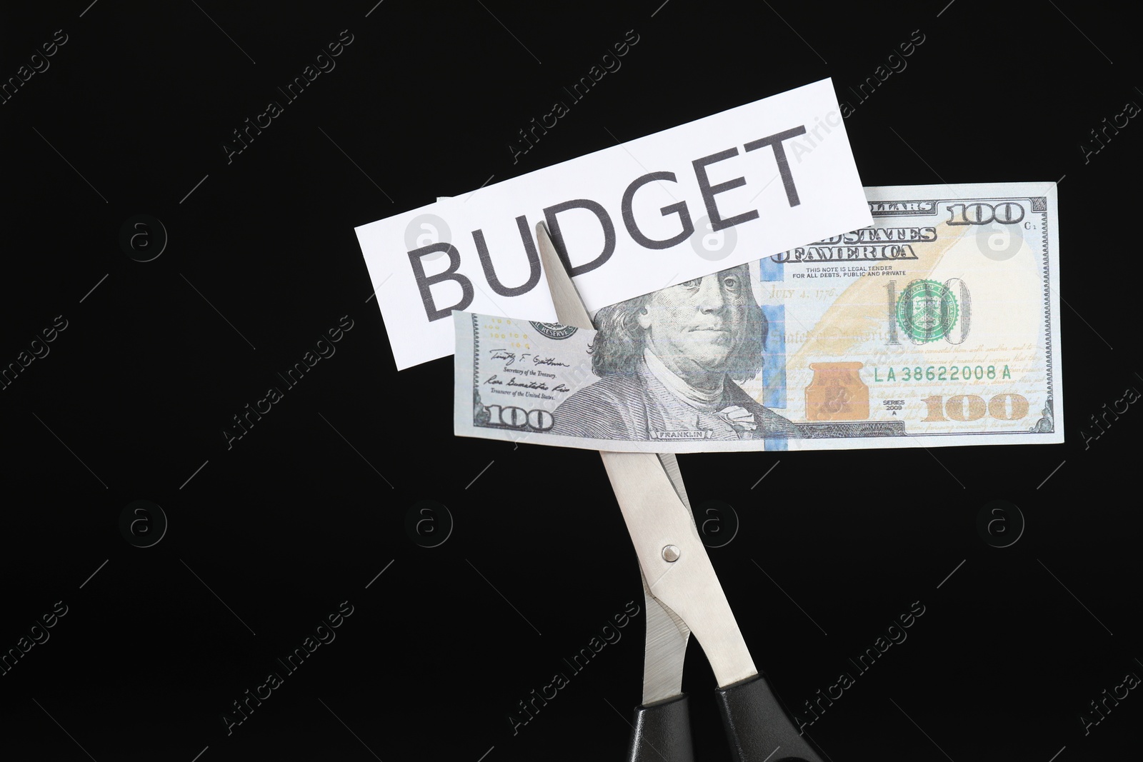 Photo of Cutting dollar banknote and paper with word Budget with scissors on black background, closeup