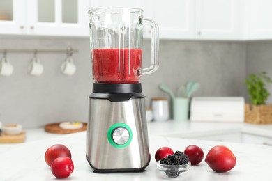 Photo of Blender with mixture of ingredients, fresh berries and fruits on white marble table indoors