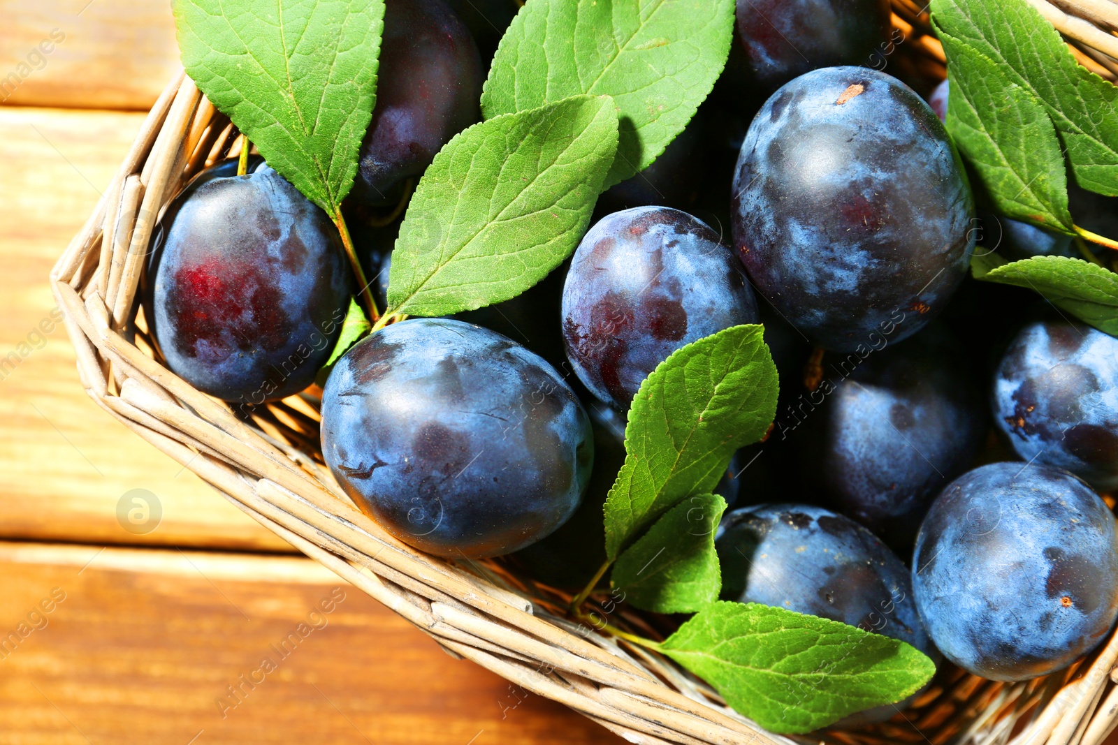 Photo of Tasty ripe plums and leaves in wicker basket on wooden table, top view