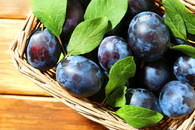 Photo of Tasty ripe plums and leaves in wicker basket on wooden table, top view