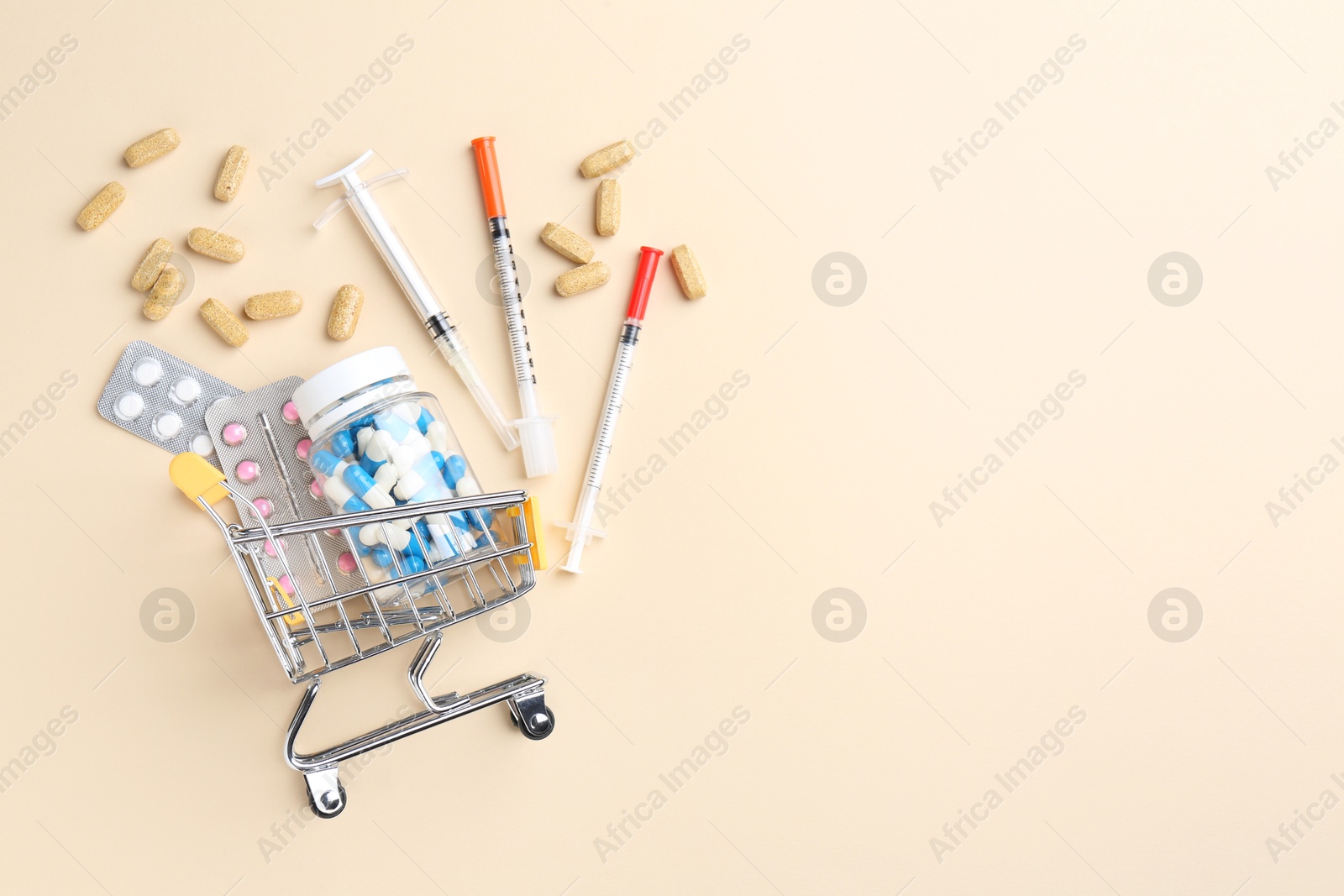 Photo of Pharmacist concept. Small shopping cart, syringes and pills on beige background, flat lay. Space for text