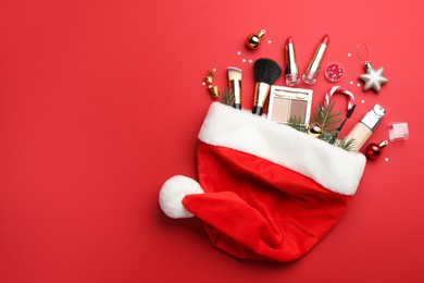 Santa hat with makeup products and Christmas decor on red background, top view