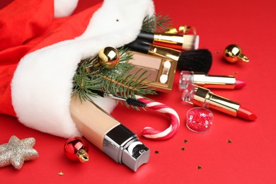 Photo of Santa hat with makeup products and Christmas decor on red background, closeup