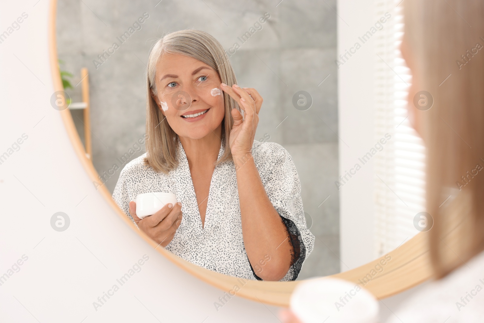 Photo of Senior woman applying face cream near mirror at home