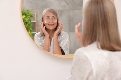 Photo of Senior woman applying face cream near mirror at home