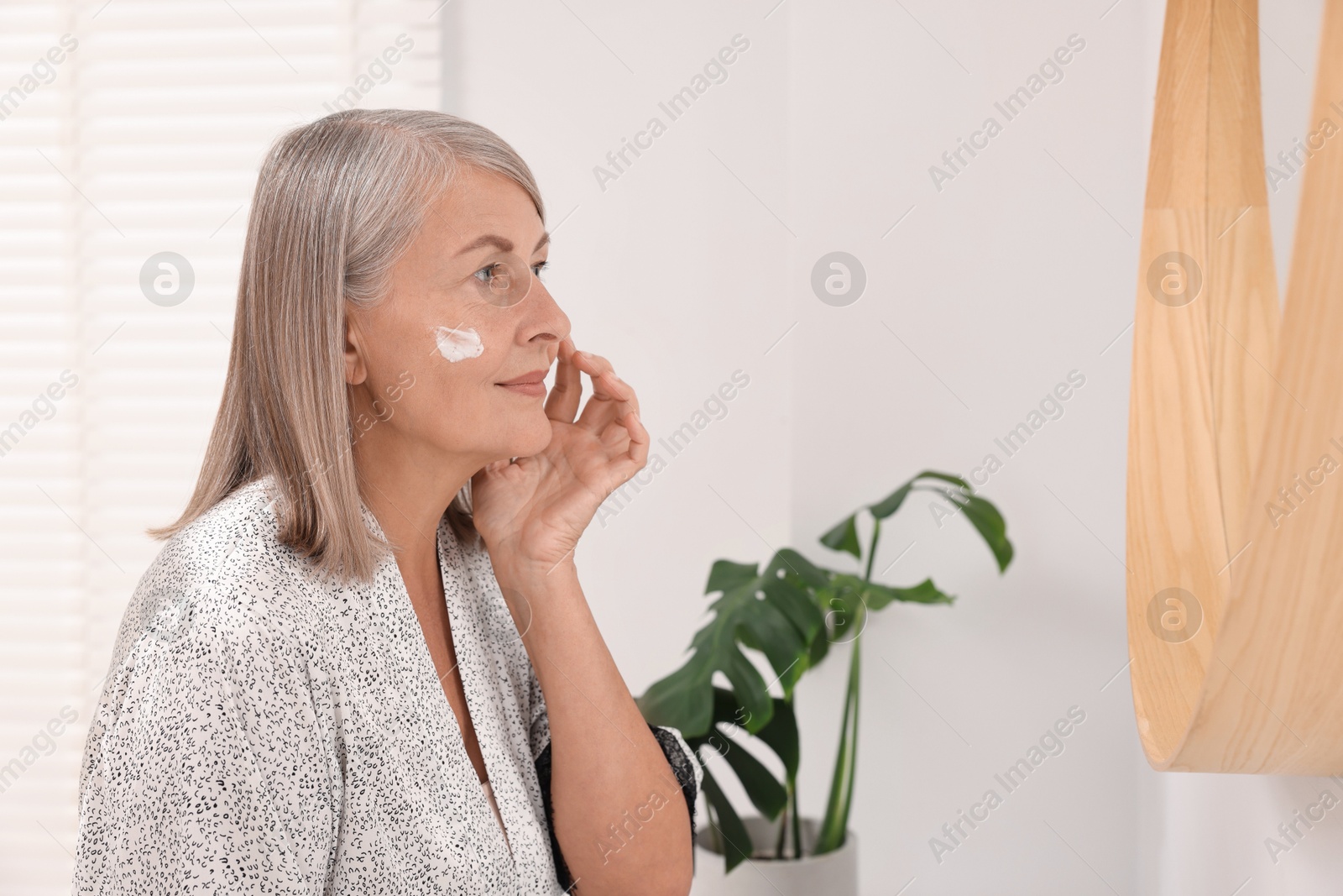 Photo of Senior woman applying face cream near mirror at home