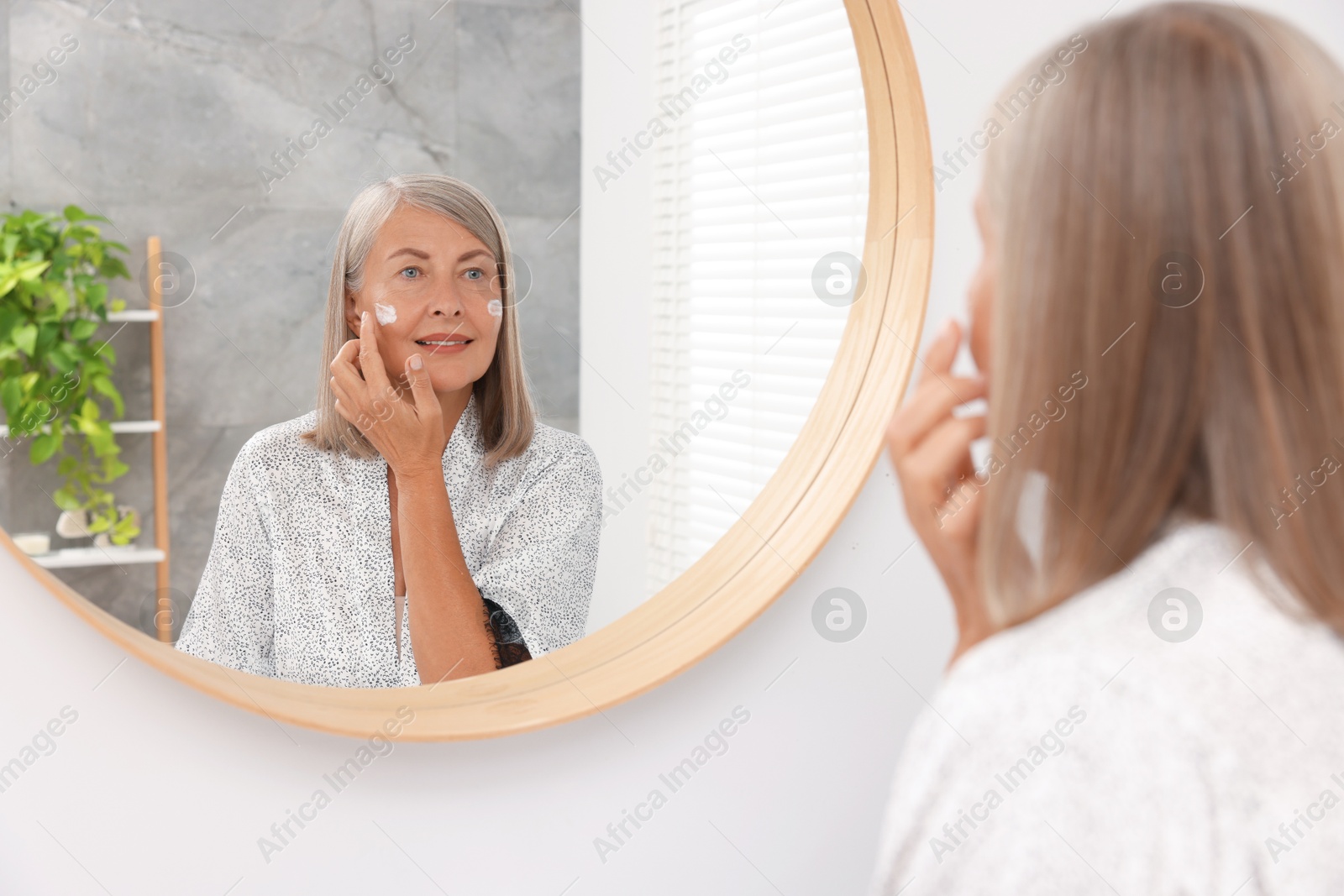 Photo of Senior woman applying face cream near mirror at home