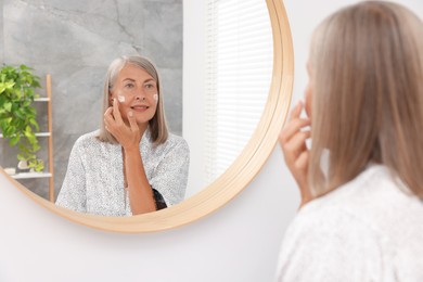 Senior woman applying face cream near mirror at home