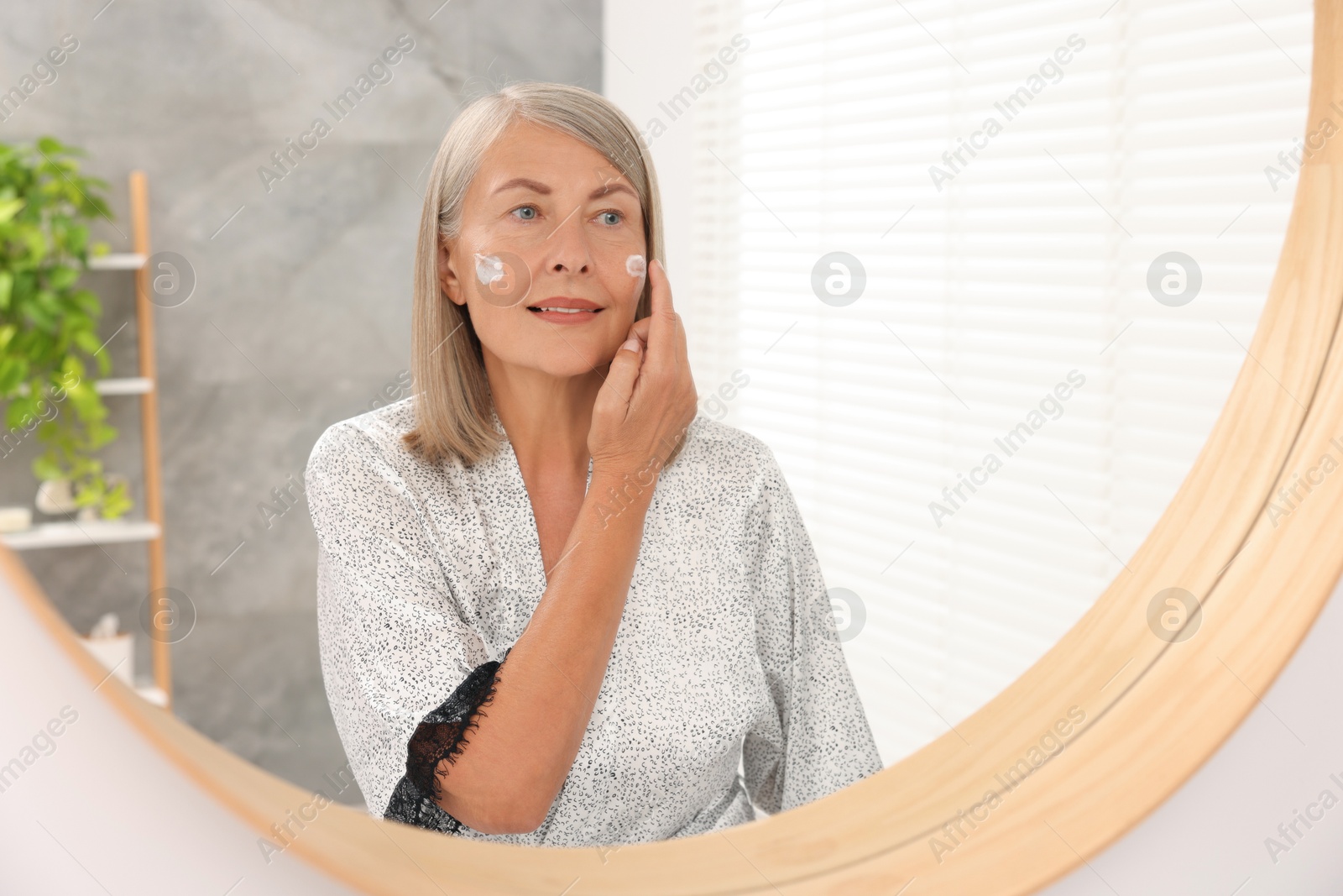 Photo of Senior woman applying face cream near mirror at home