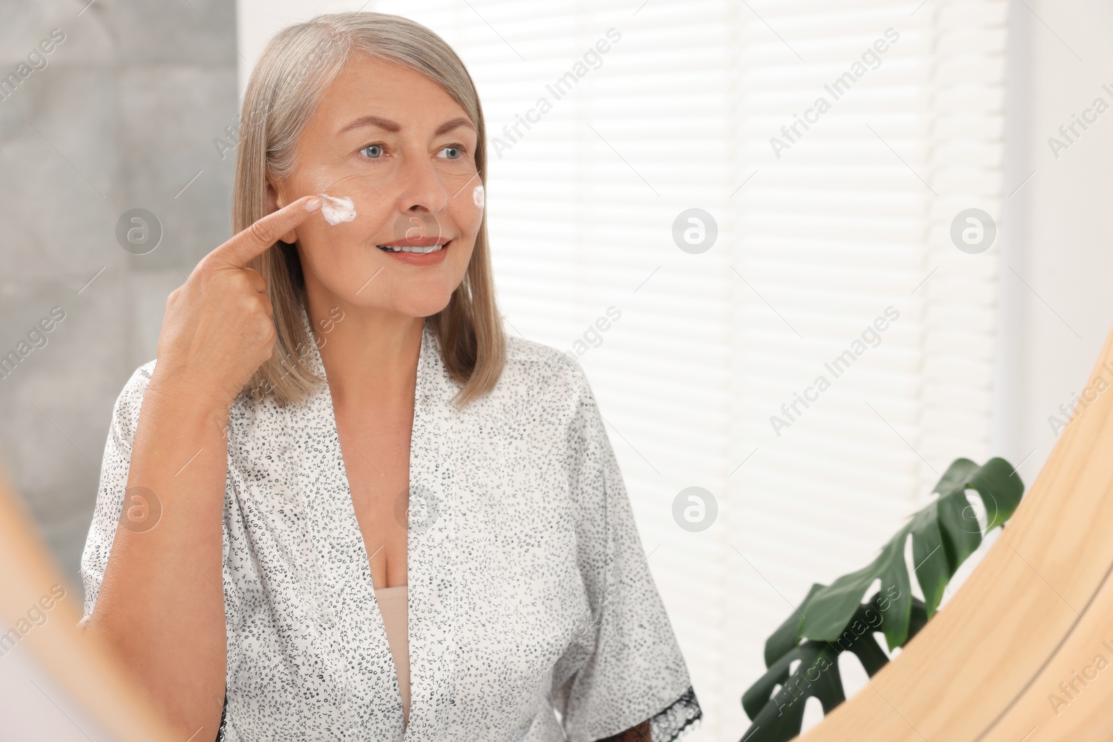 Photo of Senior woman applying face cream near mirror at home