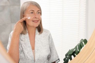 Senior woman applying face cream near mirror at home