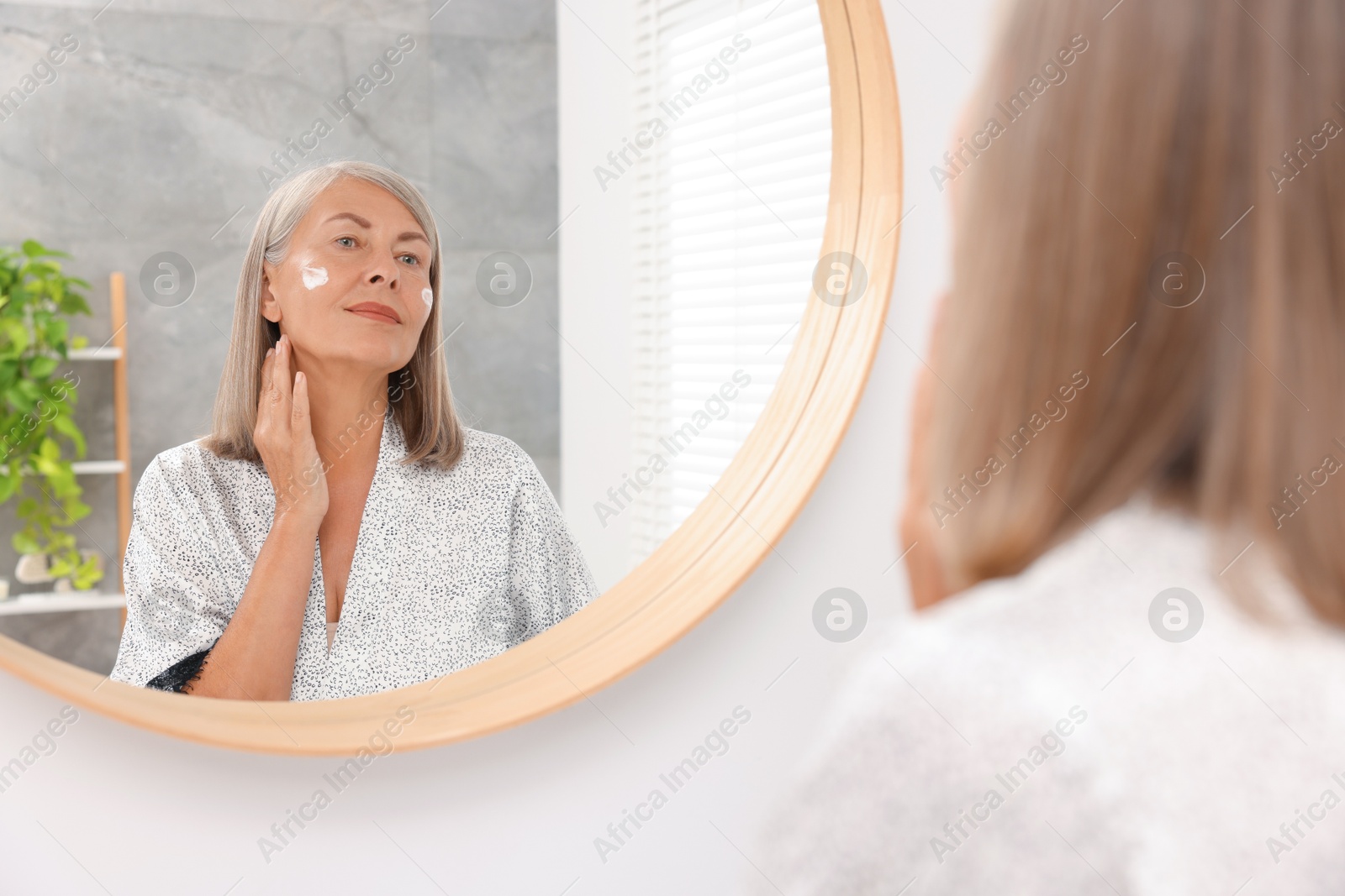 Photo of Senior woman applying face cream near mirror at home