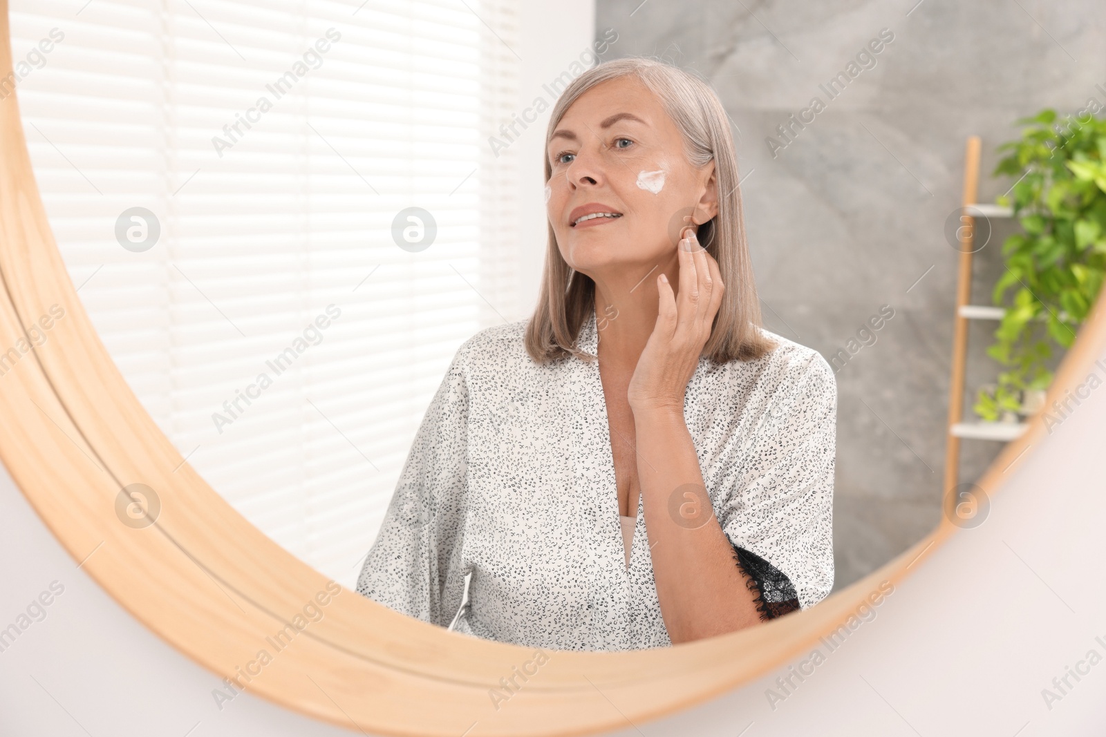 Photo of Senior woman applying face cream near mirror at home