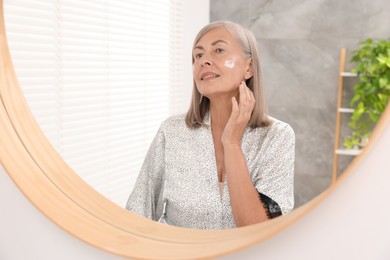 Senior woman applying face cream near mirror at home