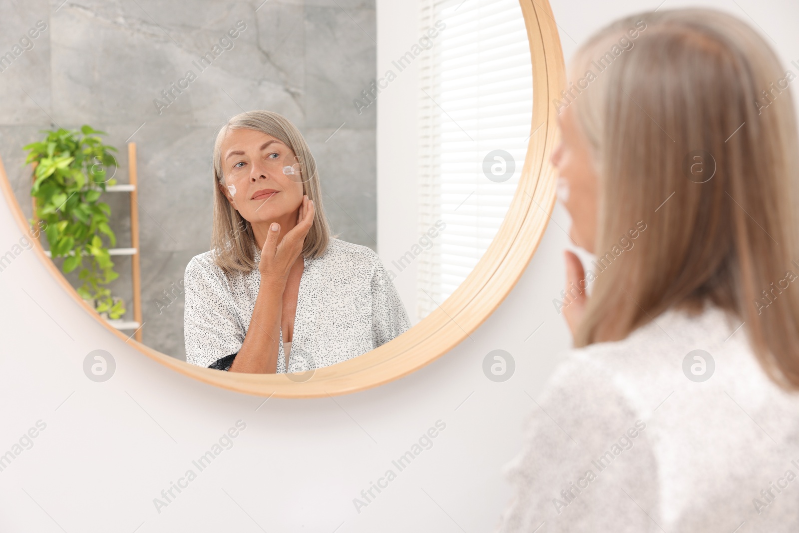 Photo of Senior woman applying face cream near mirror at home