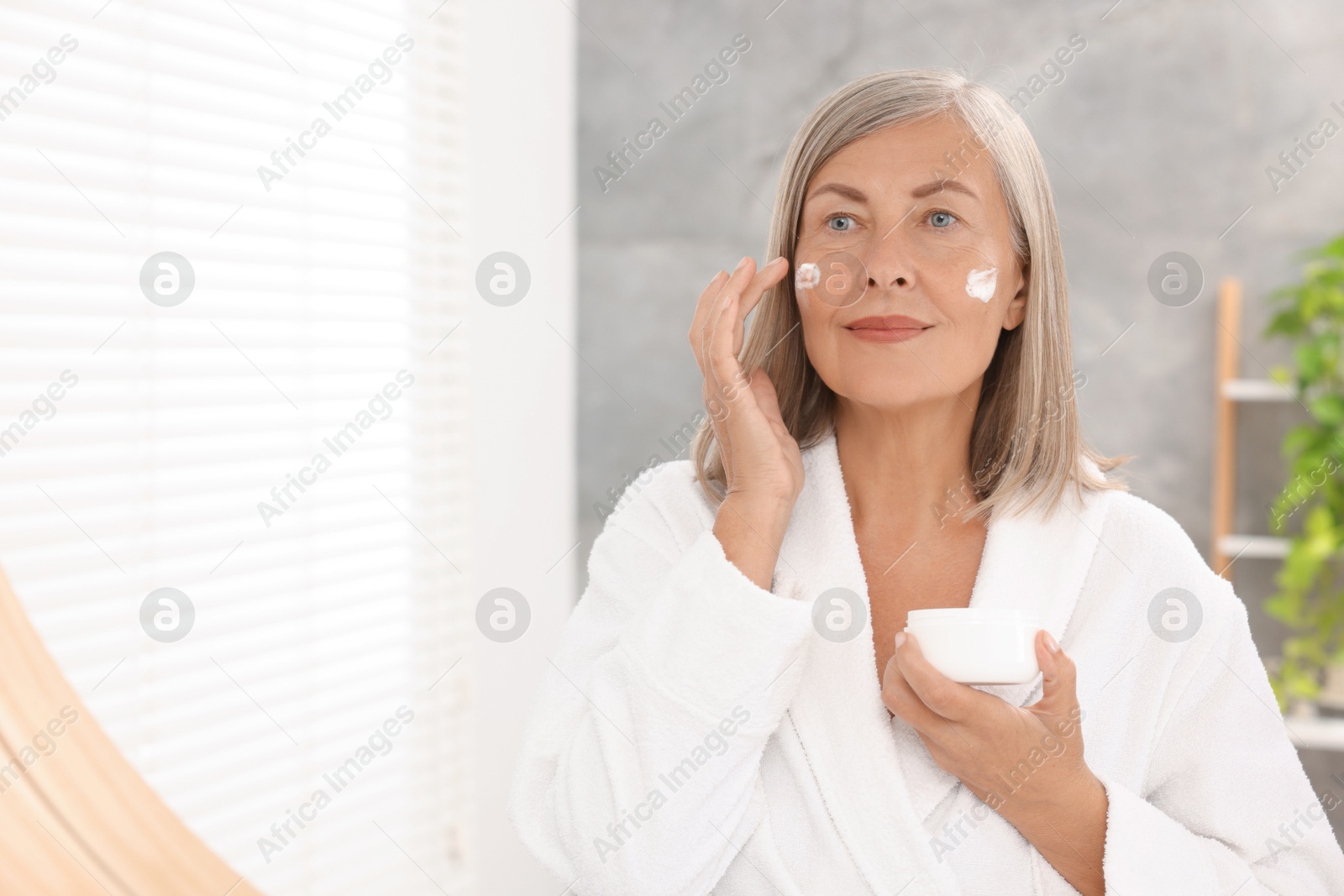 Photo of Senior woman applying face cream near mirror at home