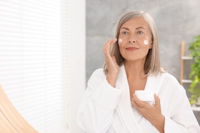 Senior woman applying face cream near mirror at home