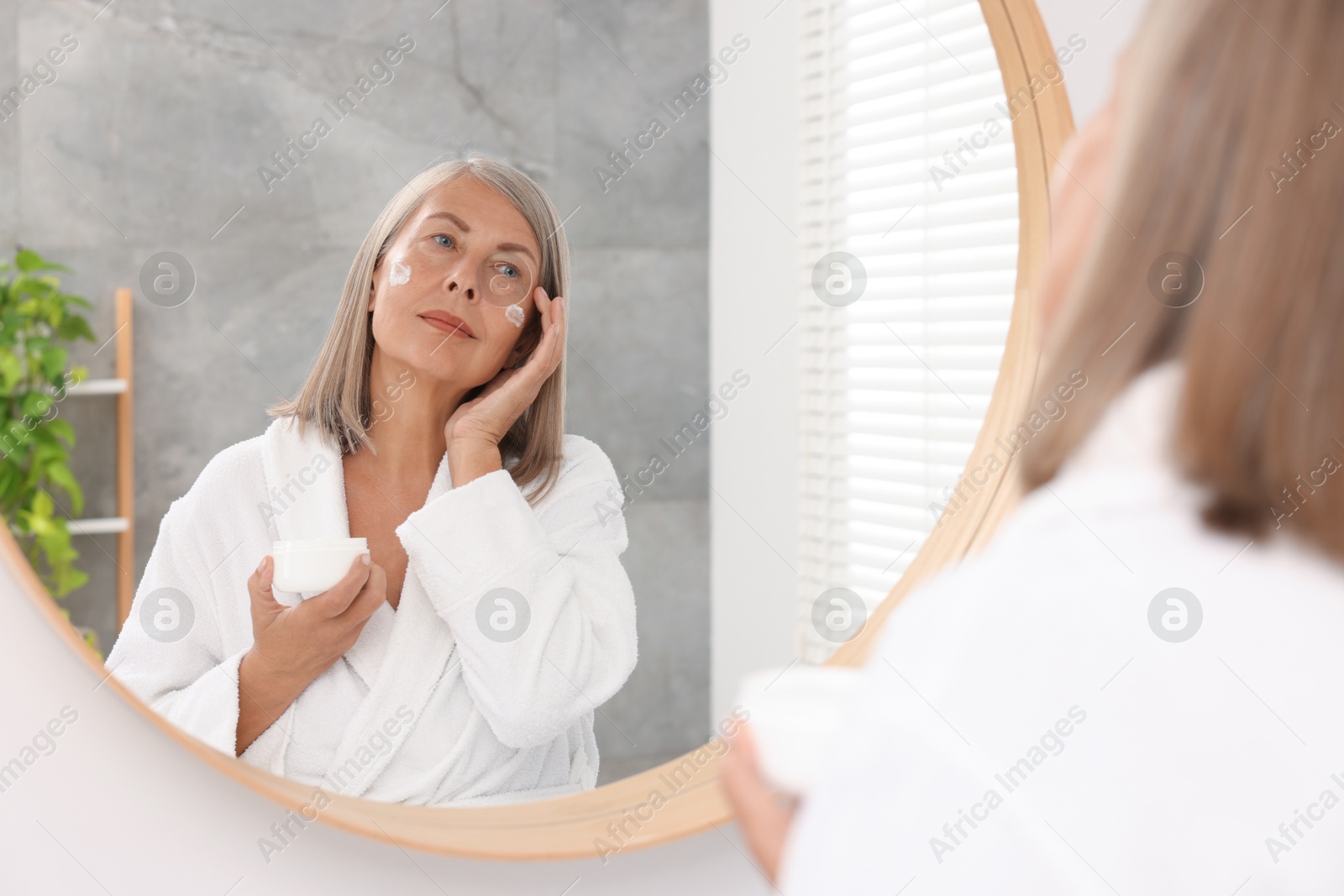 Photo of Senior woman applying face cream near mirror at home