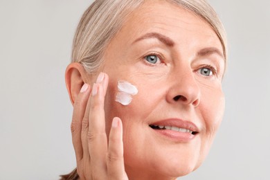 Senior woman applying face cream on light background, closeup