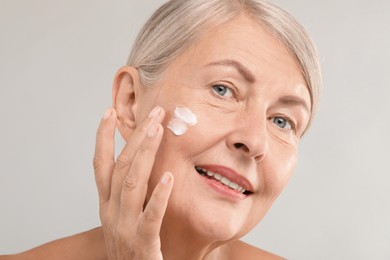 Senior woman applying face cream on light background, closeup