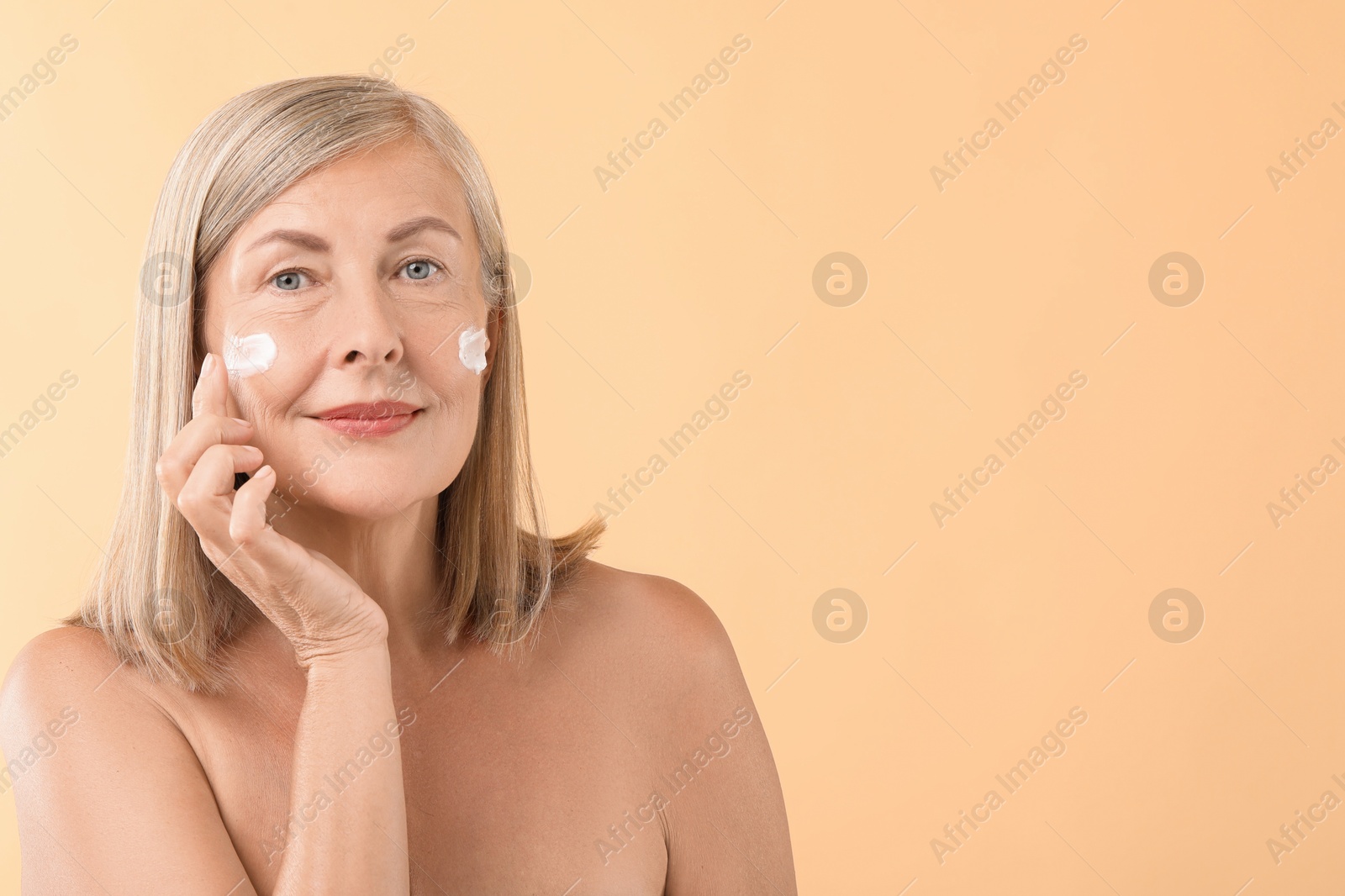 Photo of Senior woman applying face cream on beige background. Space for text