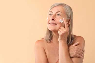 Photo of Senior woman applying face cream on beige background. Space for text