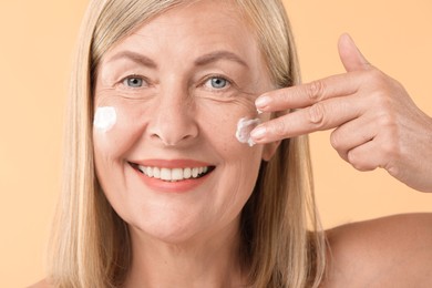 Photo of Senior woman applying face cream on beige background, closeup