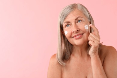 Senior woman applying face cream on pink background. Space for text