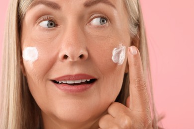 Senior woman applying face cream on pink background, closeup