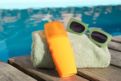 Photo of Stylish sunglasses, towel and sunscreen on wooden deck near outdoor swimming pool