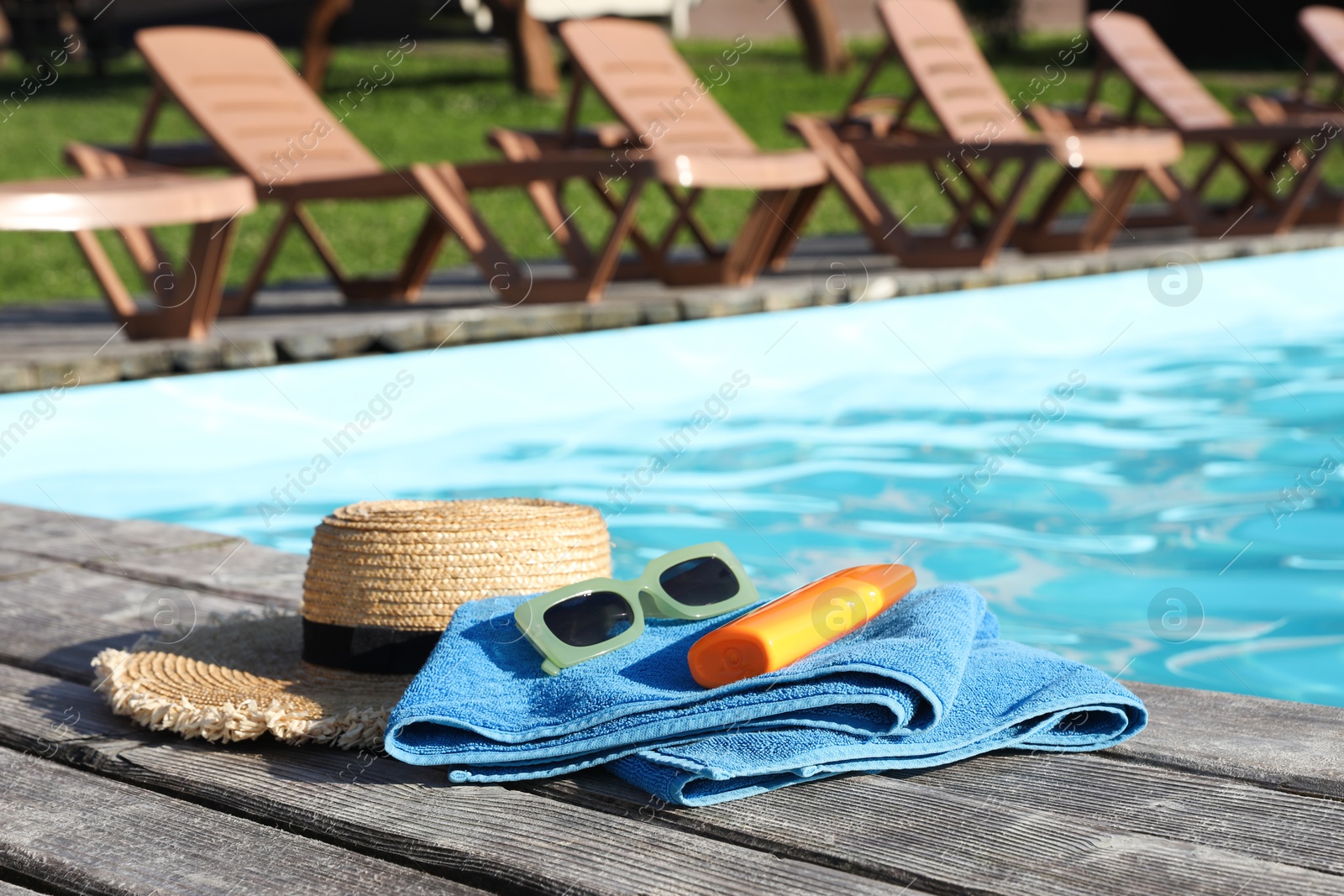 Photo of Stylish sunglasses, towel, hat and sunscreen on wooden deck near outdoor swimming pool