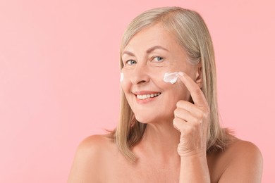 Senior woman applying face cream on pink background. Space for text