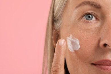 Senior woman applying face cream on pink background, closeup. Space for text