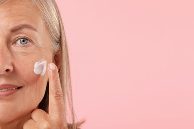 Photo of Senior woman applying face cream on pink background, closeup. Space for text