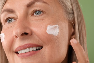 Senior woman applying face cream on green background, closeup