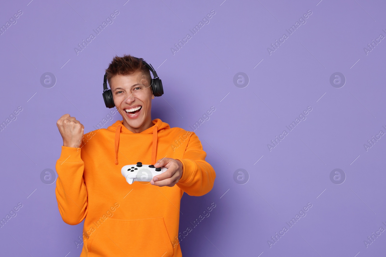 Photo of Happy young man in headphones with controller on violet background, space for text