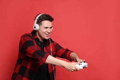 Happy young man in headphones playing video games with controller on red background, space for text