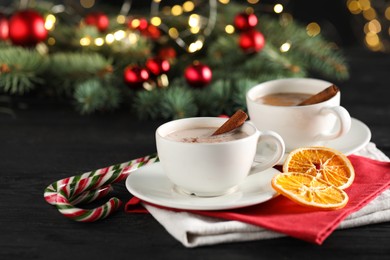 Photo of Tasty hot cocoa drink with cinnamon sticks in cups, dry orange and candy canes on black wooden table