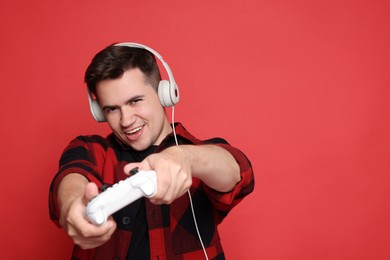 Happy young man in headphones playing video games with controller on red background, space for text