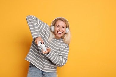 Happy woman in headphones playing video games with controller on orange background, space for text