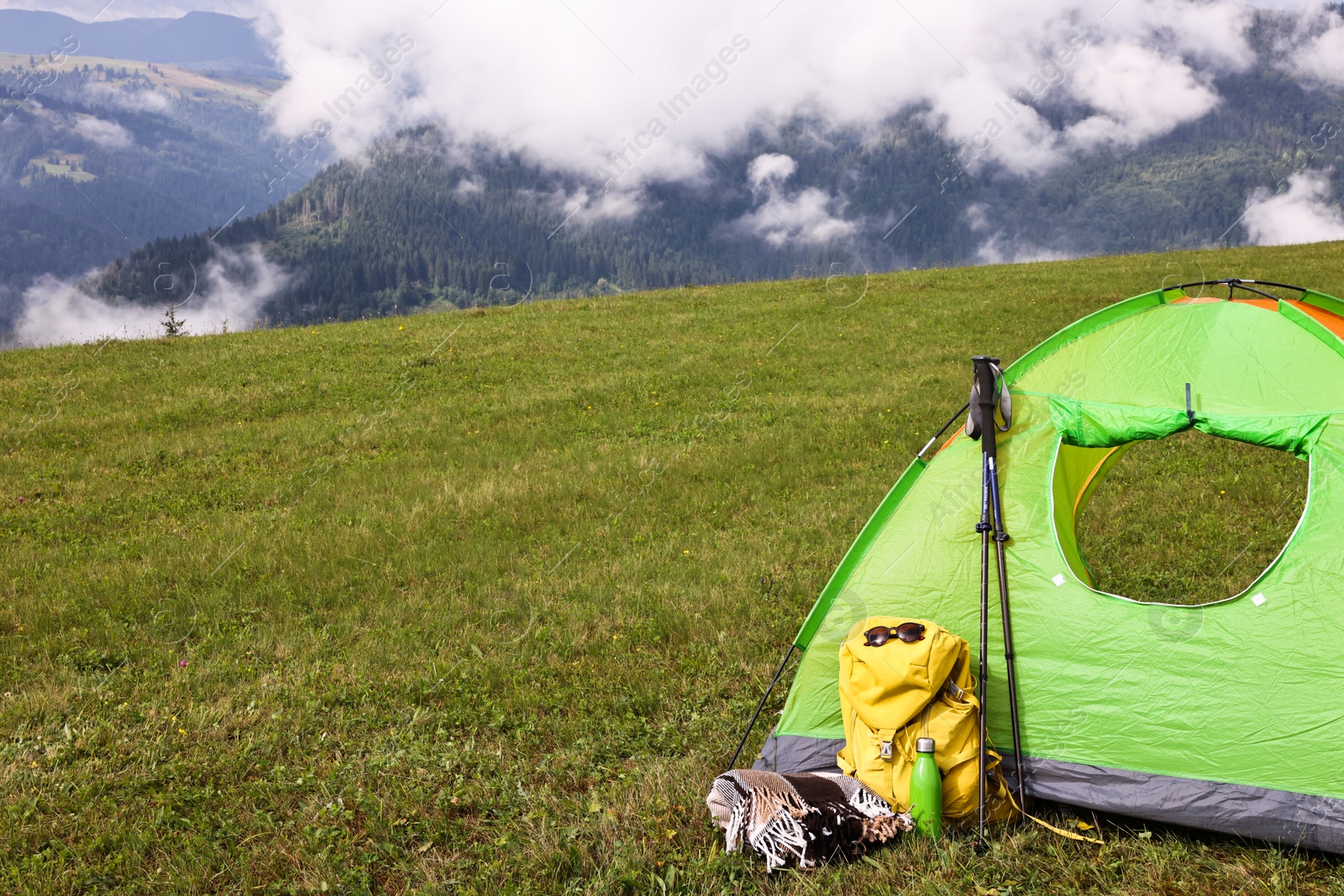 Photo of Tent, backpack, trekking poles and thermos on green grass in mountains, space for text